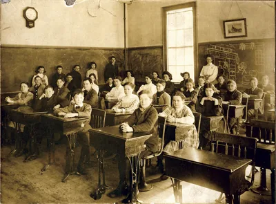 An old sepia photo of a full classroom with a teacher and rows of students at wooden desks