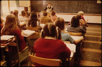 Students at desks facing a teacher at the blackboard in a classroom