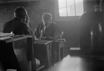 A black and white photo of a teacher at a desk in an old classroom by a tall window