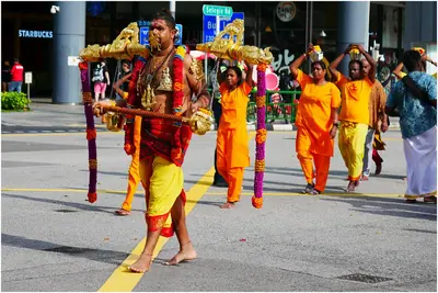 A devotee in yellow carries a decorated kavadi on his shoulders down a street, others in orange follow
