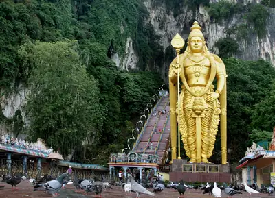 A golden statue of Murugan holding a spear beside long steps to a cave in a green hill