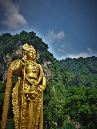 The giant golden statue of Lord Murugan holding his vel, against green hills and blue sky