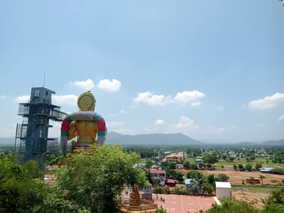 A golden Murugan statue on a hilltop with fields and hills behind