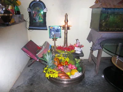 A bronze vessel with fruits, yellow konna flowers and coconut before a tall lit brass lamp and a picture of Krishna
